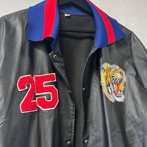 May include: A black jacket featuring a blue and red striped collar, a red felt number 25 patch on the right chest, and an embroidered tiger head patch on the left chest. The jacket has a front snap button closure and a smooth, dark finish.