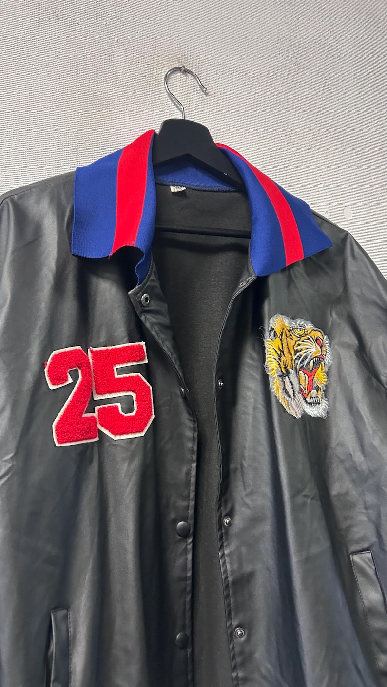 May include: A black jacket featuring a blue and red striped collar, a red felt number 25 patch on the right chest, and an embroidered tiger head patch on the left chest. The jacket has a front snap button closure and a smooth, dark finish.