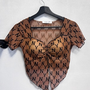 May include: Brown mesh top with black "NY" pattern. The top has short sleeves, a sweetheart neckline, and a ruched center. The top is hanging on a black hanger.