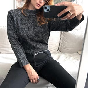 May include: A dark grey, long-sleeved jumper with a high neckline. The jumper has a textured, shimmering appearance. Paired with black jeans and a thin black belt. The person is holding a black phone.