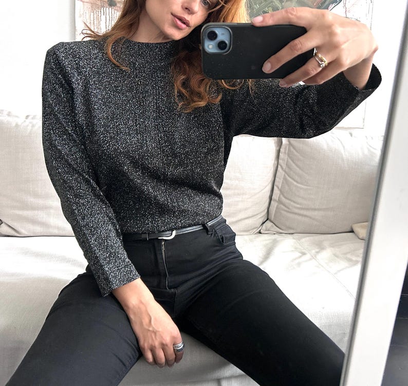 May include: A dark grey, long-sleeved jumper with a high neckline. The jumper has a textured, shimmering appearance. Paired with black jeans and a thin black belt. The person is holding a black phone.