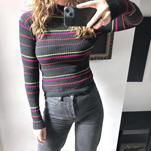 Könnte beinhalten: Schwarzes Rippstrick-Langarm-Top mit horizontalen Streifen in Pink, Gelb und Rot. Das Top hat einen Rollkragen und wird mit einer dunkelgrauen Jeans kombiniert. Die Person hält ein schwarzes Smartphone.