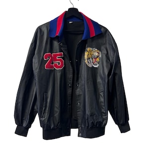 May include: A black jacket featuring a red number 25 patch on the right chest and an embroidered tiger head patch on the left chest. The jacket has a blue and red collar, long sleeves with ribbed cuffs, a ribbed hem, and a front snap button closure.