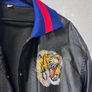 May include: A black satin jacket featuring a vibrant embroidered tiger head patch on the chest. The jacket has a fold-over collar in bright blue with a red stripe detail, a front snap-button closure, and a smooth, slightly reflective fabric finish.