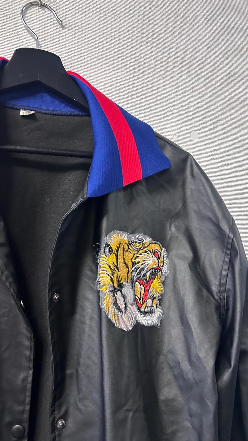 May include: A black satin jacket featuring a vibrant embroidered tiger head patch on the chest. The jacket has a fold-over collar in bright blue with a red stripe detail, a front snap-button closure, and a smooth, slightly reflective fabric finish.