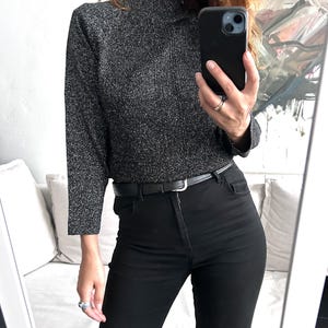 May include: A black and silver metallic mock neck top with long sleeves, paired with black high-waisted jeans and a black belt. The outfit is completed with silver rings. The image is taken in front of a mirror.