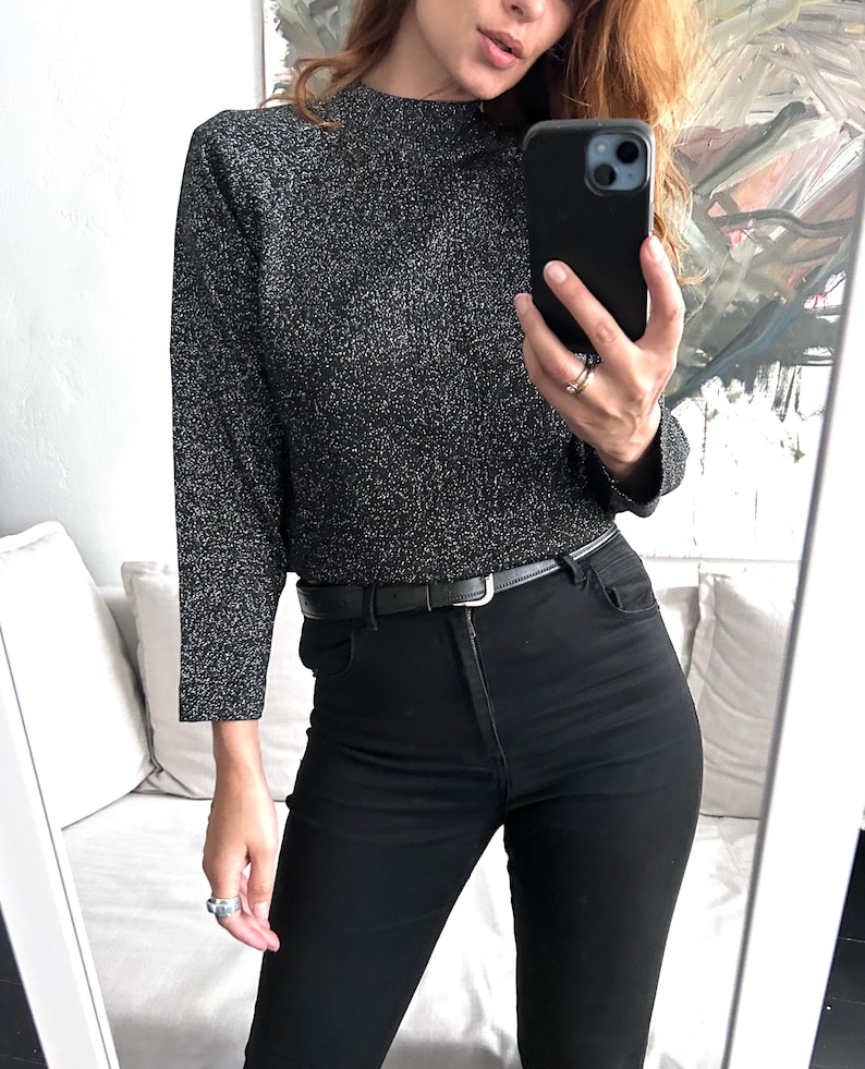 May include: A black and silver metallic mock neck top with long sleeves, paired with black high-waisted jeans and a black belt. The outfit is completed with silver rings. The image is taken in front of a mirror.