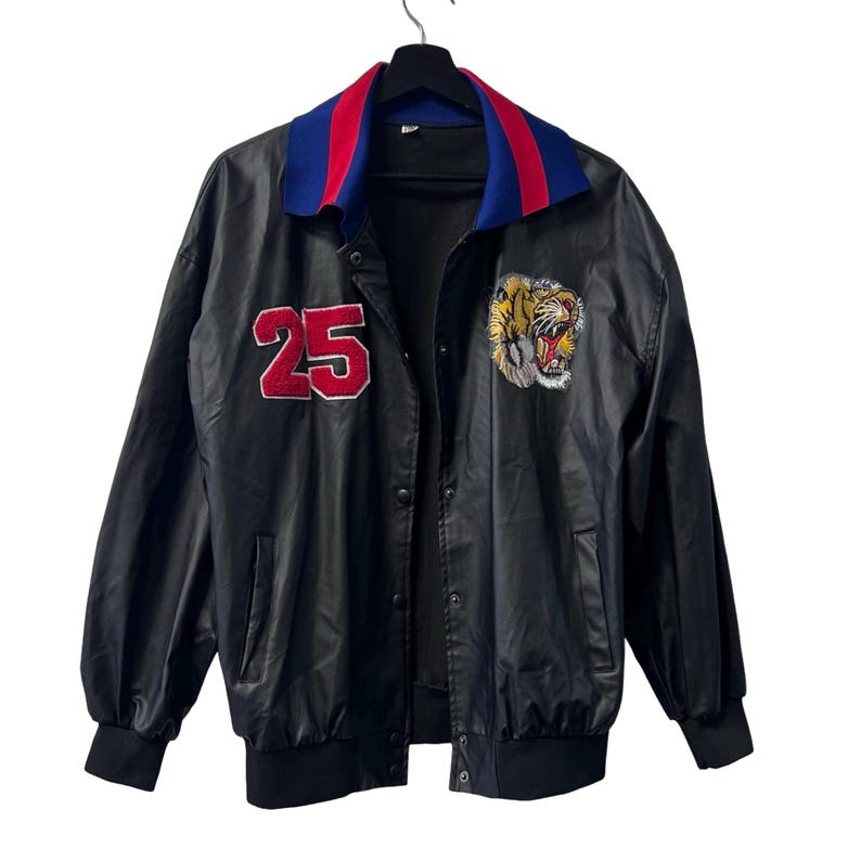 May include: A black jacket featuring a red number 25 patch on the right chest and an embroidered tiger head patch on the left chest. The jacket has a blue and red collar, long sleeves with ribbed cuffs, a ribbed hem, and a front snap button closure.