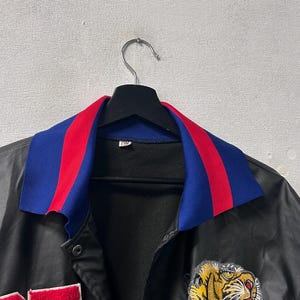 May include: A black satin-finish jacket featuring a blue and red striped collar, a large embroidered tiger head patch on the chest, and the number 25 in red with white trim on the side. The jacket has a black interior lining and metal snap buttons.