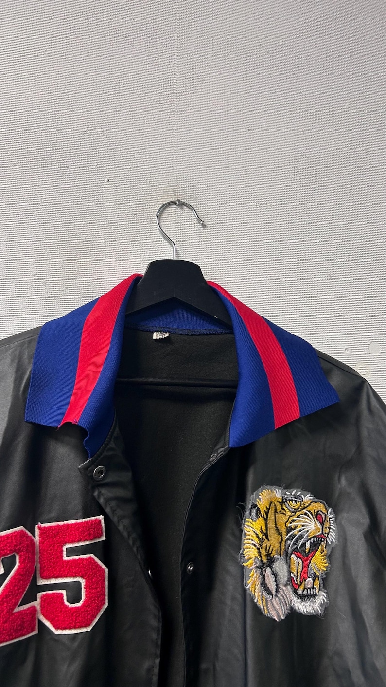 May include: A black satin-finish jacket featuring a blue and red striped collar, a large embroidered tiger head patch on the chest, and the number 25 in red with white trim on the side. The jacket has a black interior lining and metal snap buttons.