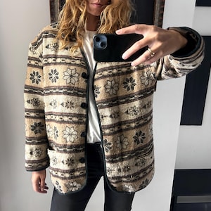Op de afbeelding: Een patroonjas in tinten bruin en beige, met zwarte afwerking en knopen. De jas heeft een bloemen- en geometrisch ontwerp. Het heeft twee voorzakken en wordt over een wit shirt gedragen.