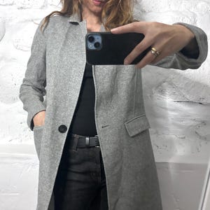 Pode incluir: Um casaco de lã cinza claro com um único botão preto, lapela entalhada e bolsos frontais. O casaco é usado sobre uma blusa preta e jeans escuros. A pessoa está segurando um telefone preto.