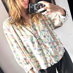 Könnte beinhalten: Eine Bluse mit floralem Muster, V-Ausschnitt und langen Ärmeln. Die Bluse hat ein Blumenmuster in Rot-, Grün- und Blautönen auf cremefarbenem Hintergrund. Die Ärmel haben geknöpfte Manschetten.