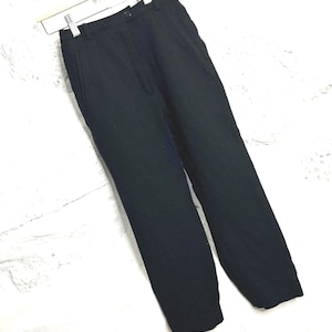 May include: Black trousers hanging on a white hanger against a white brick wall. The pants have a button closure and pockets. The fabric appears to be a woven material. The pants are straight-legged.