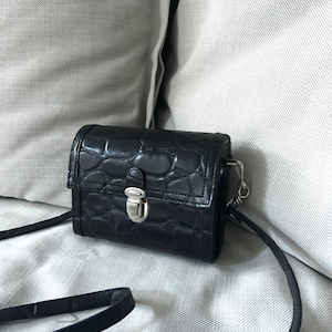 Puede incluir: Un pequeño bolso bandolera de cuero negro con cierre plateado y textura de piel de cocodrilo.