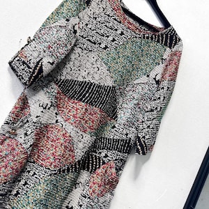 Pode incluir: Um vestido multicolorido em estilo patchwork com mangas curtas. O vestido apresenta um decote redondo e um tecido texturizado com uma mistura de cores, incluindo preto, branco, vermelho e verde. O vestido está pendurado em um cabide preto.