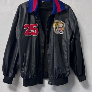 May include: A black jacket featuring a red number 25 patch on the right chest and a tiger head embroidery on the left chest. The jacket has a blue and red collar, long sleeves with black cuffs, a snap button front closure, and two side pockets.