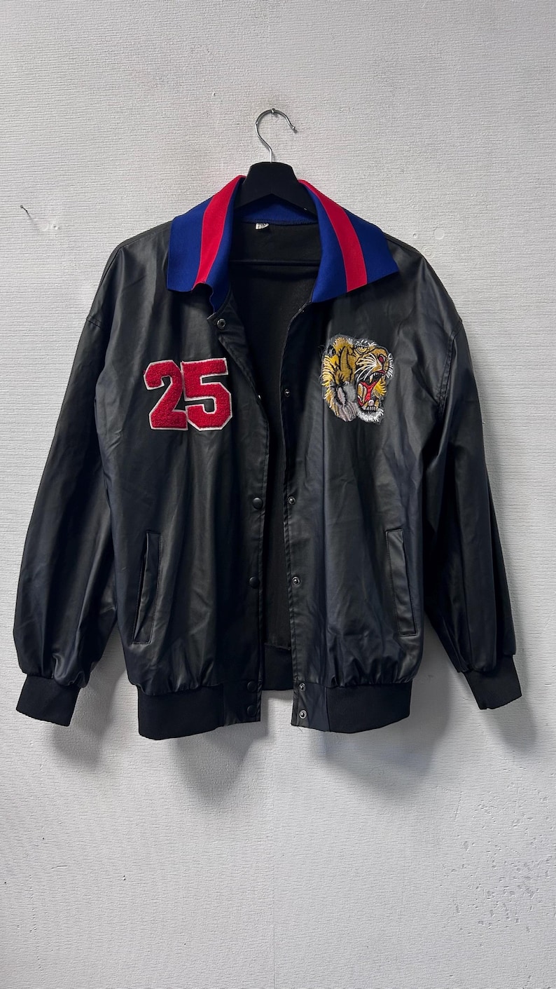 May include: A black jacket featuring a red number 25 patch on the right chest and a tiger head embroidery on the left chest. The jacket has a blue and red collar, long sleeves with black cuffs, a snap button front closure, and two side pockets.