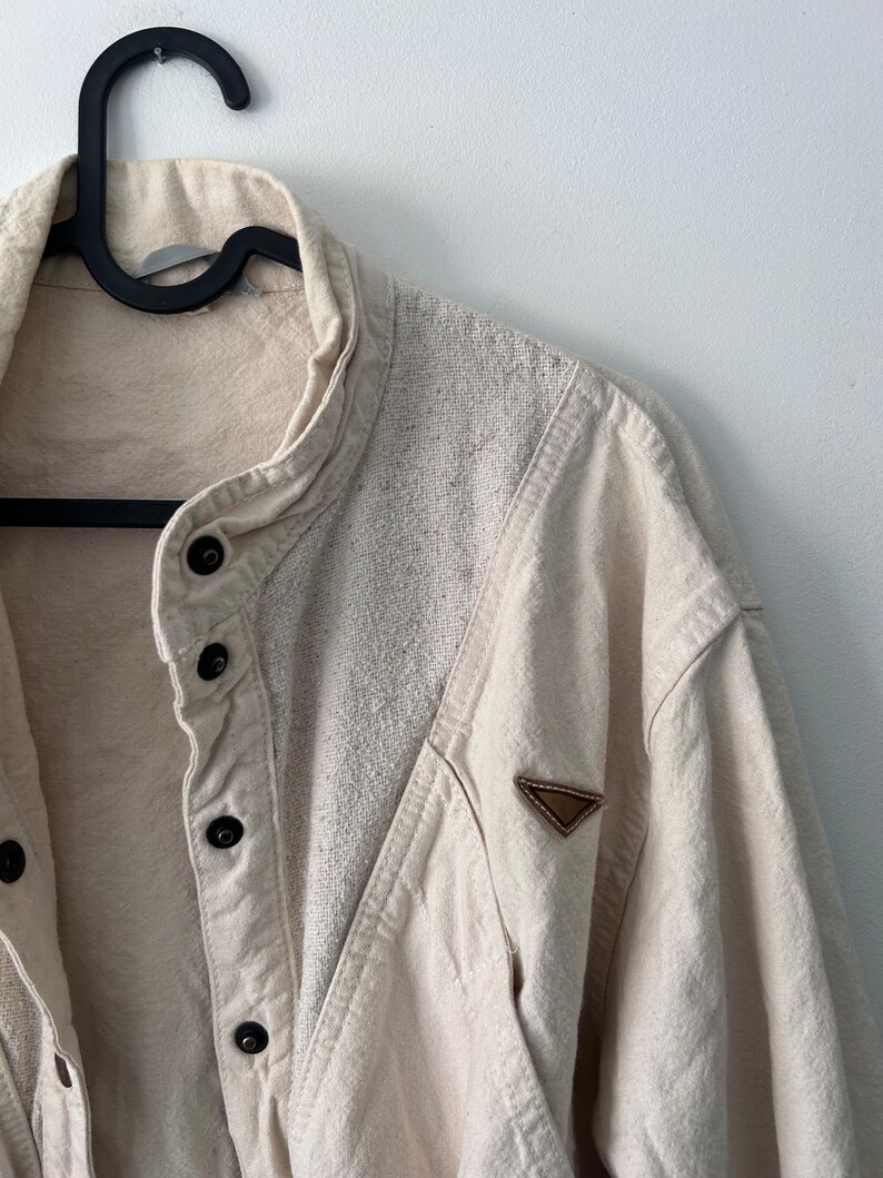 K&ouml;nnte beinhalten: Ein cremefarbenes Hemd mit Kn&ouml;pfen und einem braunen Leder-Patch am linken &Auml;rmel. Das Hemd hat einen Stehkragen und einen Knopfverschluss.