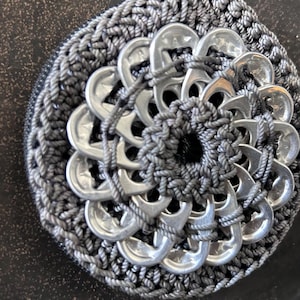 Op de afbeelding: Een ronde portemonnee gemaakt van donkergrijs gehaakt garen, met een decoratieve inleg in het midden van in elkaar grijpende zilverkleurige aluminium bliklipjes in een radiaal patroon, met een zwarte ritssluiting langs de buitenrand.