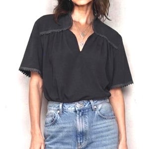 Op de afbeelding: Een zwarte blouse met korte mouwen, een split-V-halslijn en decoratieve randen langs de schouders en mouwboorden. Het kledingstuk wordt gecombineerd met een lichtblauwe spijkerbroek met een hoge taille en zichtbare metalen knopen.