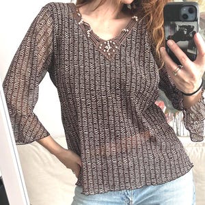 Puede incluir: Una blusa marrón transparente con un estampado geométrico blanco. La blusa tiene mangas largas con puños acampanados y un escote en V adornado con ganchillo y pequeñas cuentas blancas. El dobladillo inferior también está fruncido.