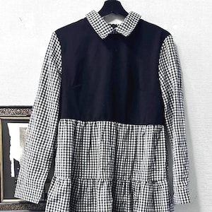 May include: A black and white gingham dress with long sleeves and a contrasting black bodice. The dress features a white and black checkered collar and a tiered skirt. The dress is hanging on a black hanger against a white wall.