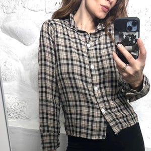 Peut inclure: Une chemise à carreaux beige, noire et blanche avec des boutons. La chemise à manches longues a un col classique et des boutons blancs. La chemise est portée avec un pantalon noir. Le fond est un mur blanc texturé.
