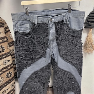 May include: Grey denim jeans with distressed black fabric detailing. The jeans have a button fastening, belt loops, and a unique design with shredded fabric. The jeans are displayed on a wooden hanger.