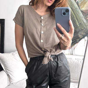 Può includere: Una camicia marrone a maniche corte con bottoni, annodata in vita, indossata con pantaloni di pelle neri.