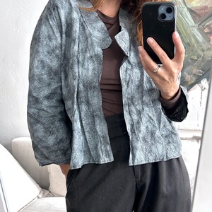 Könnte beinhalten: Eine langärmelige Jacke mit einem gesprenkelten, grau-schwarzen Texturmuster, offenem Vorderteil mit umgeschlagenem Kragen und lockerer Passform. Die Jacke wird über einem einfarbigen braunen Oberteil getragen und mit einer schwarzen Hose kombiniert.