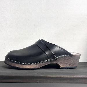 May include: A black leather clog with a smooth finish and a wide strap across the top. The shoe features a thick, natural-toned wooden sole with visible white stitching connecting the leather upper to the wood base and a dark rubber tread.