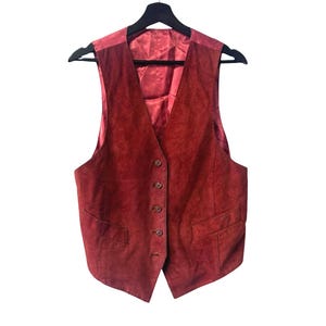 May include: A burgundy suede vest with a five-button fastening and two front pockets hangs on a black hanger. The vest has a satin lining and a classic, tailored design.