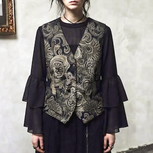 Può includere: Un gilet nero con un dettagliato motivo paisley color oro e verde oliva, indossato sopra un top nero a maniche lunghe con volant a campana. Il gilet presenta uno scollo a V e una chiusura frontale con bottoni, con maniche dal design stratificato.