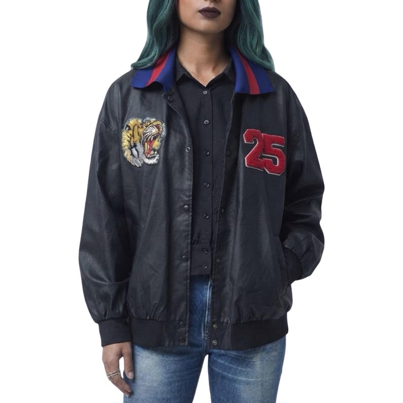May include: A black jacket with a snap-button front, featuring a blue and red striped collar. The chest displays an embroidered tiger head patch on the right side and a large red number 25 patch on the left side, with black ribbed cuffs.