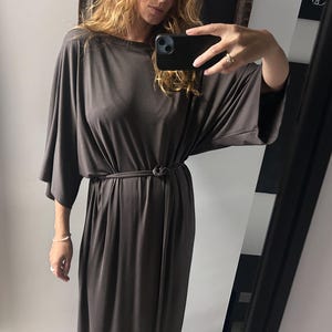 Pode incluir: Um vestido maxi cinza escuro com decote barco e mangas largas. O vestido é acinturado na cintura com um cinto de tecido a condizer. O vestido é feito de um material macio e fluido, adequado para uso casual.