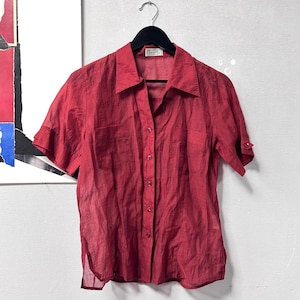 May include: A red button-down shirt with short sleeves, hanging on a black hanger. The shirt has two chest pockets and a collar. The fabric appears lightweight and slightly sheer. The shirt is a solid, deep red colour.