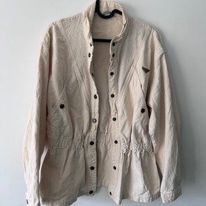 K&ouml;nnte beinhalten: Eine cremefarbene, lang&auml;rmelige Jacke mit Knopfleiste und Kordelzug in der Taille. Die Jacke hat einen Stehkragen und einen Aufn&auml;her am rechten &Auml;rmel.