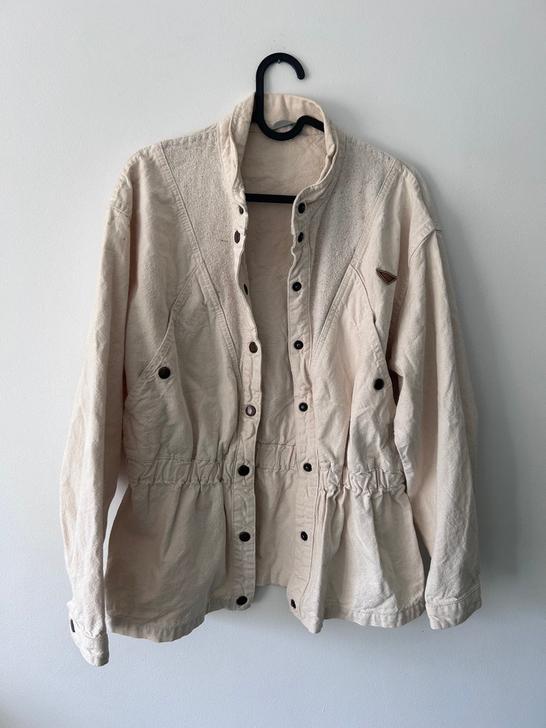 K&ouml;nnte beinhalten: Eine cremefarbene, lang&auml;rmelige Jacke mit Knopfleiste und Kordelzug in der Taille. Die Jacke hat einen Stehkragen und einen Aufn&auml;her am rechten &Auml;rmel.
