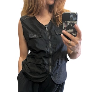 Può includere: Gilet nero senza maniche con scollo a V, cerniera frontale e tasche multiple. Il gilet ha un aspetto leggermente lucido e una piccola etichetta sulla tasca sinistra del petto. Il gilet è indossato sopra un completo nero.
