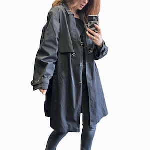 Könnte beinhalten: Ein schwarzer Trenchcoat mit klassischem Design. Der Mantel hat eine doppelreihige Front mit großen Knöpfen, einen Reverskragen und eine taillierte Taille. Die Ärmel haben geknöpfte Manschetten. Der Mantel ist knielang und aus glattem, wasserabweisendem Stoff.