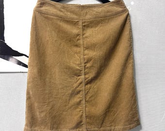 May include: A tan corduroy skirt hanging on a black hanger. The skirt has a straight cut and a visible seam down the back. The fabric has vertical ridges, and the skirt appears to be made of a medium-weight material.