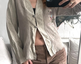 May include: A light beige linen button-up shirt with long sleeves and a floral embroidery design on the right side. The shirt is open at the front, revealing a glimpse of the wearer's midriff.