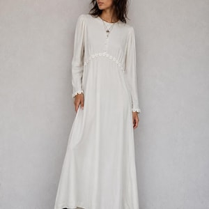 May include: A floor-length, long-sleeved off-white dress featuring a round neckline, a button-down bodice, and delicate white floral lace trim detailing along the waistline and sleeve cuffs, paired with black ankle boots.