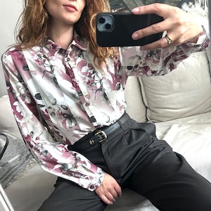 Könnte beinhalten: Eine Frau trägt ein langärmeliges, weißes und rosafarbenes Hemd mit Blumendruck und Knöpfen, einen schwarzen Gürtel und eine schwarze Hose.