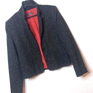 Puede incluir: Un blazer gris oscuro con un patrón moteado. El blazer tiene un forro interior rojo y un cuello con muescas. Cuenta con dos botones para cerrar y mangas largas. La chaqueta está colgada en una percha negra.