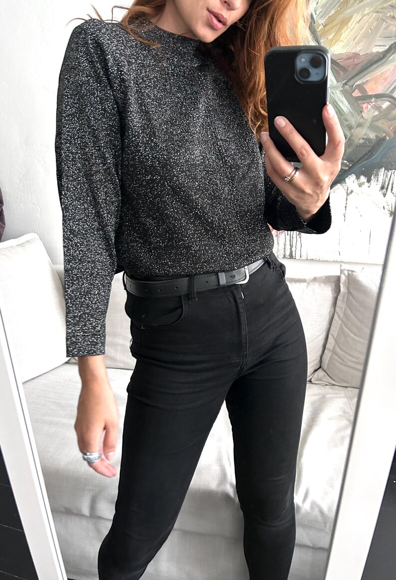 May include: A long-sleeved, dark grey, sparkly jumper paired with black jeans and a black belt. The outfit is reflected in a mirror, showcasing a casual, everyday style.