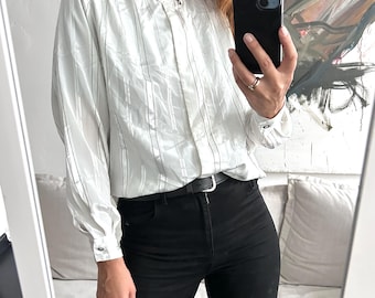 May include: A white long-sleeve shirt with black vertical stripes and a high collar. The shirt has a button closure at the neck and a single button on the collar. The shirt is worn with black pants and a black belt.