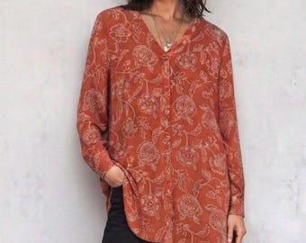 May include: A long-sleeved button-down shirt in a burnt orange hue featuring a repeating white floral line-art pattern. The shirt has a V-neckline, a straight hem, and a relaxed fit, paired with dark charcoal pants to complete the ensemble.