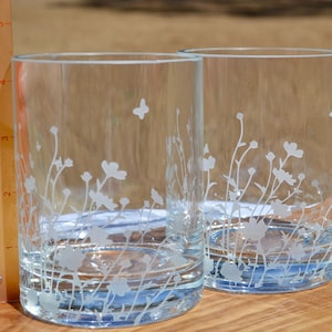 May include: Two clear glass tumblers with an etched design of white wildflowers and a butterfly. The glasses have a blue tint at the bottom.